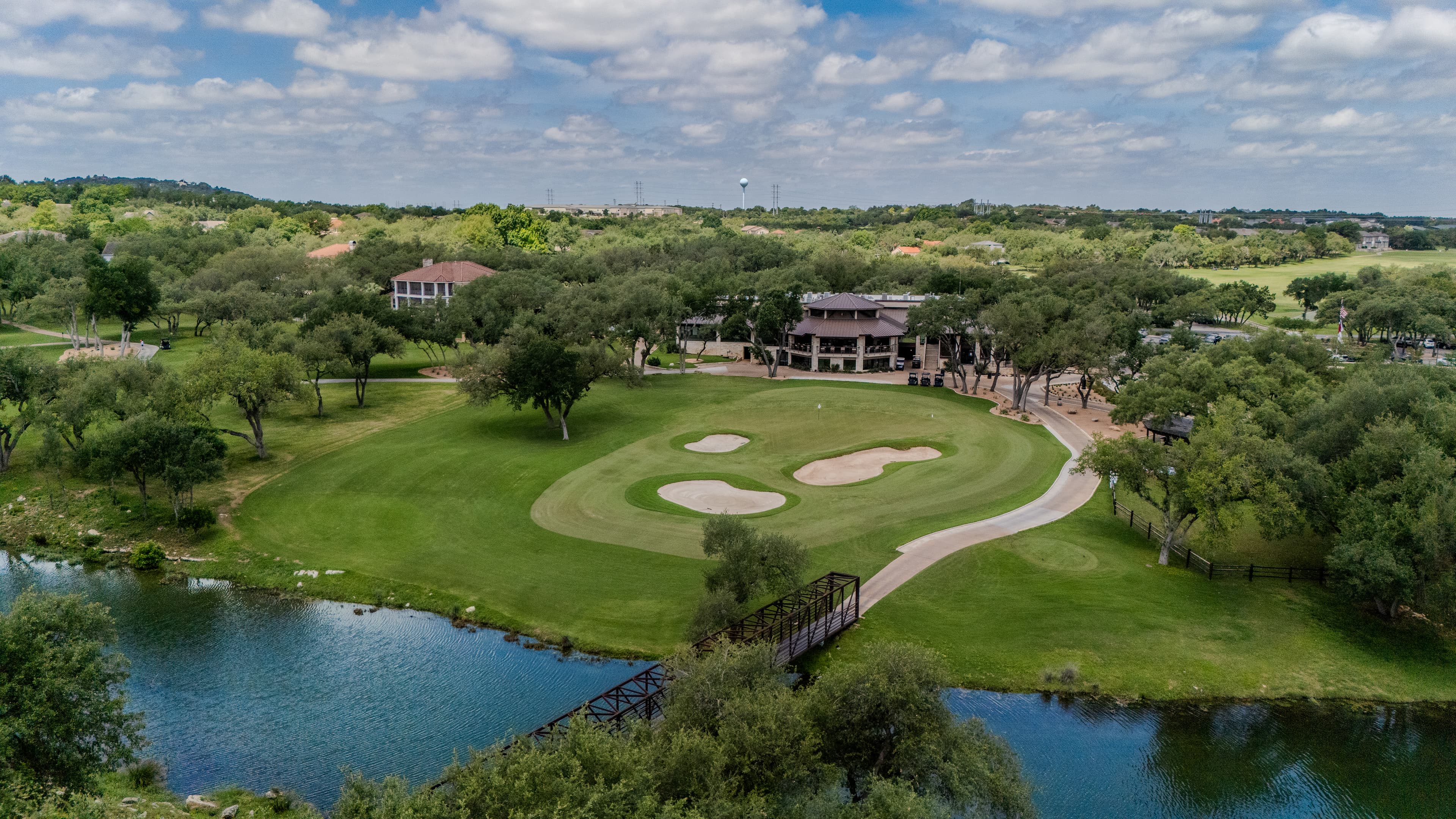Lush green golf course with sand bunkers, a bridge over water, and a clubhouse.