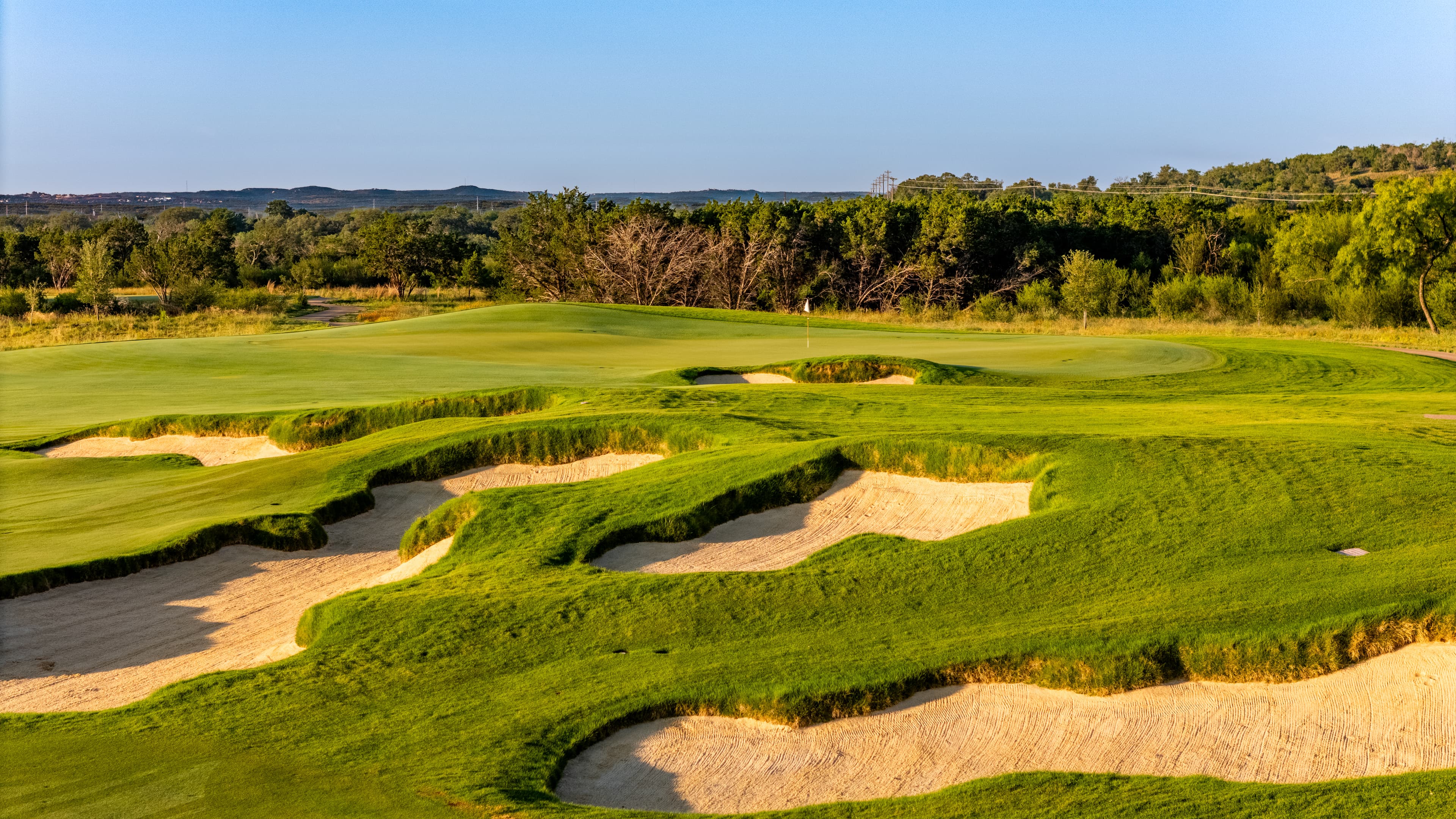 Sunlit golf course with undulating green fairways, deep sand bunkers, and a distant flagstick.