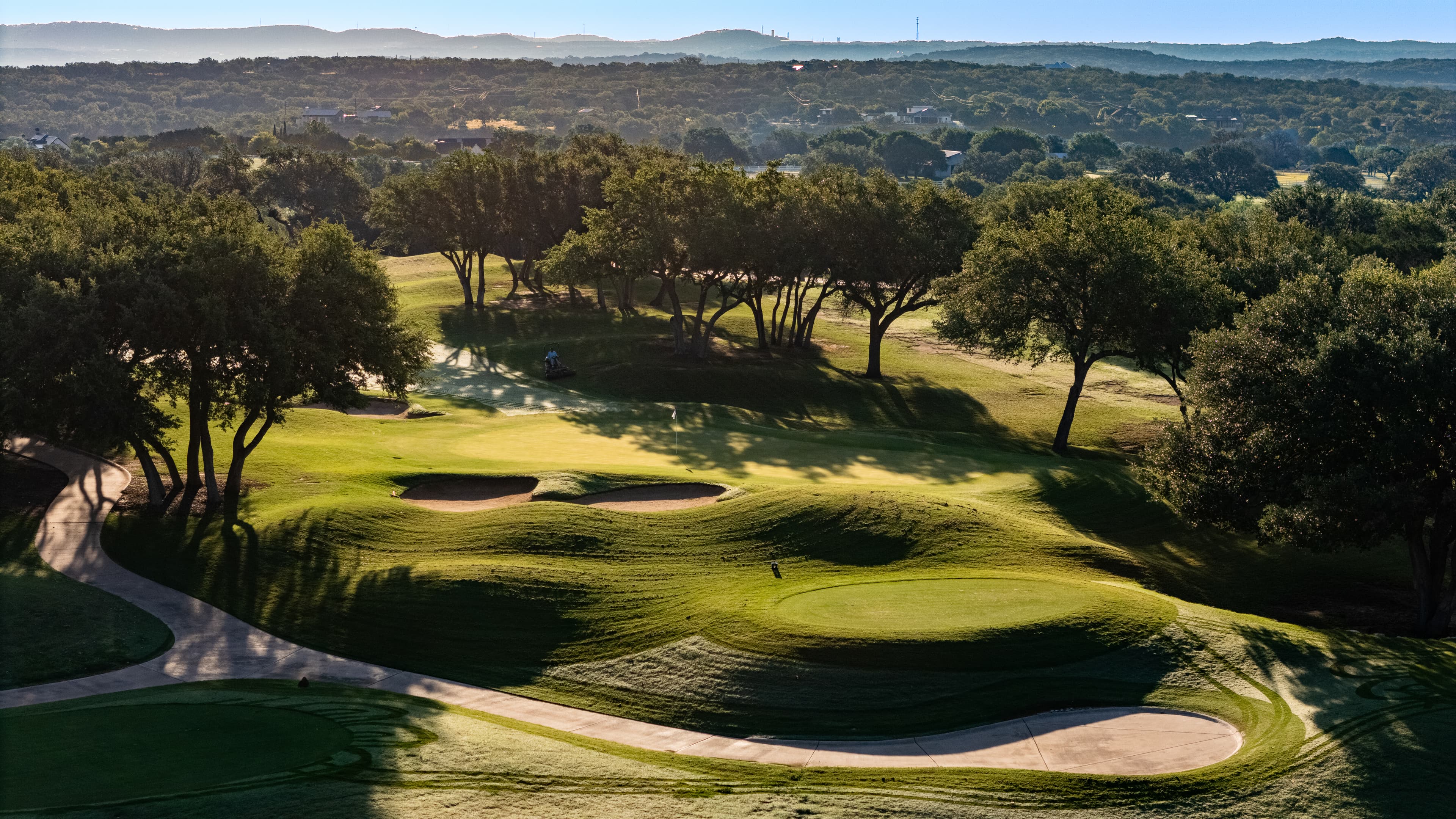 Sunlit golf course featuring rolling green hills, sand traps, and trees casting long shadows.