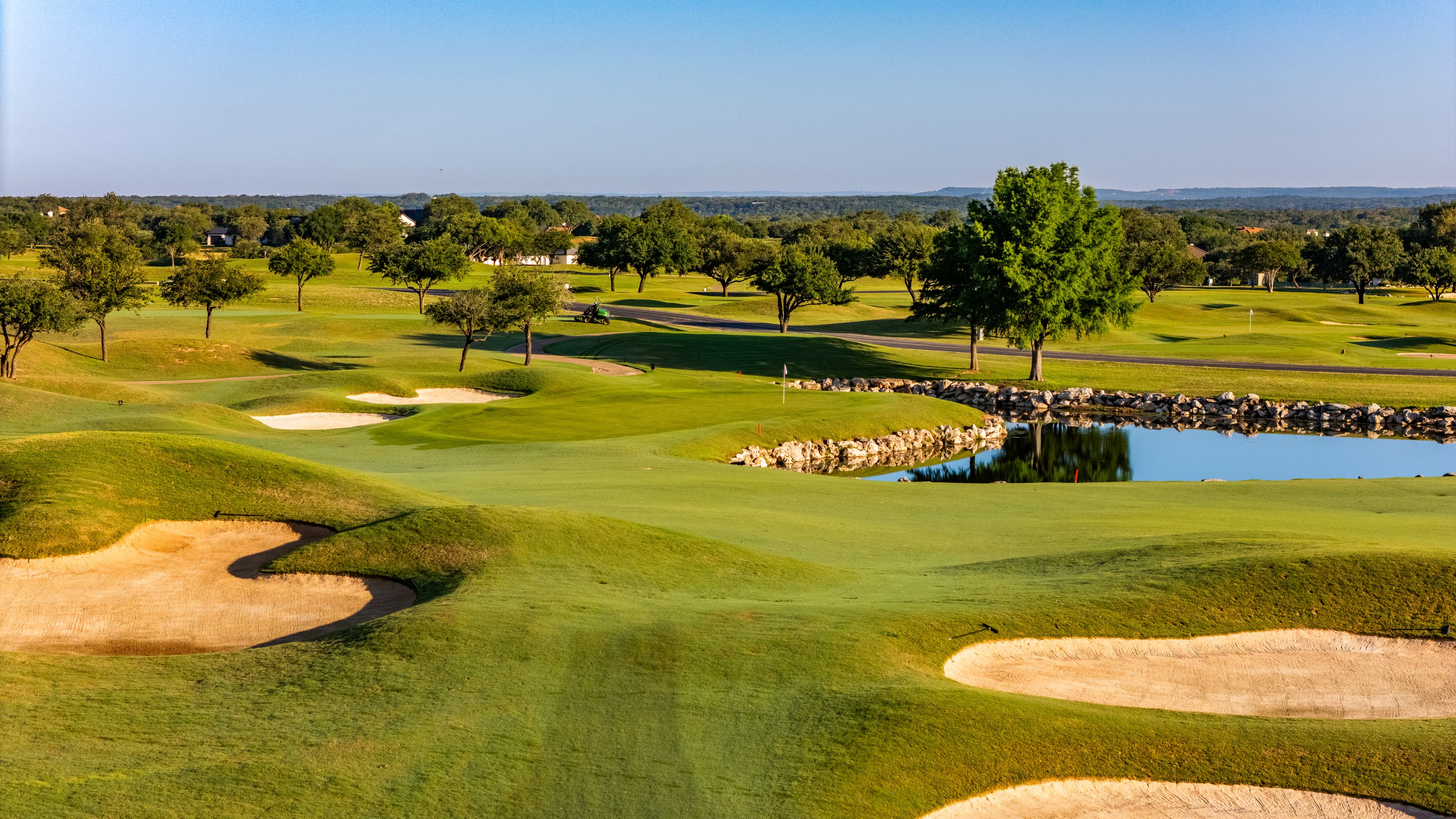 Rolling green golf course fairways with sand bunkers and a small pond under clear skies.