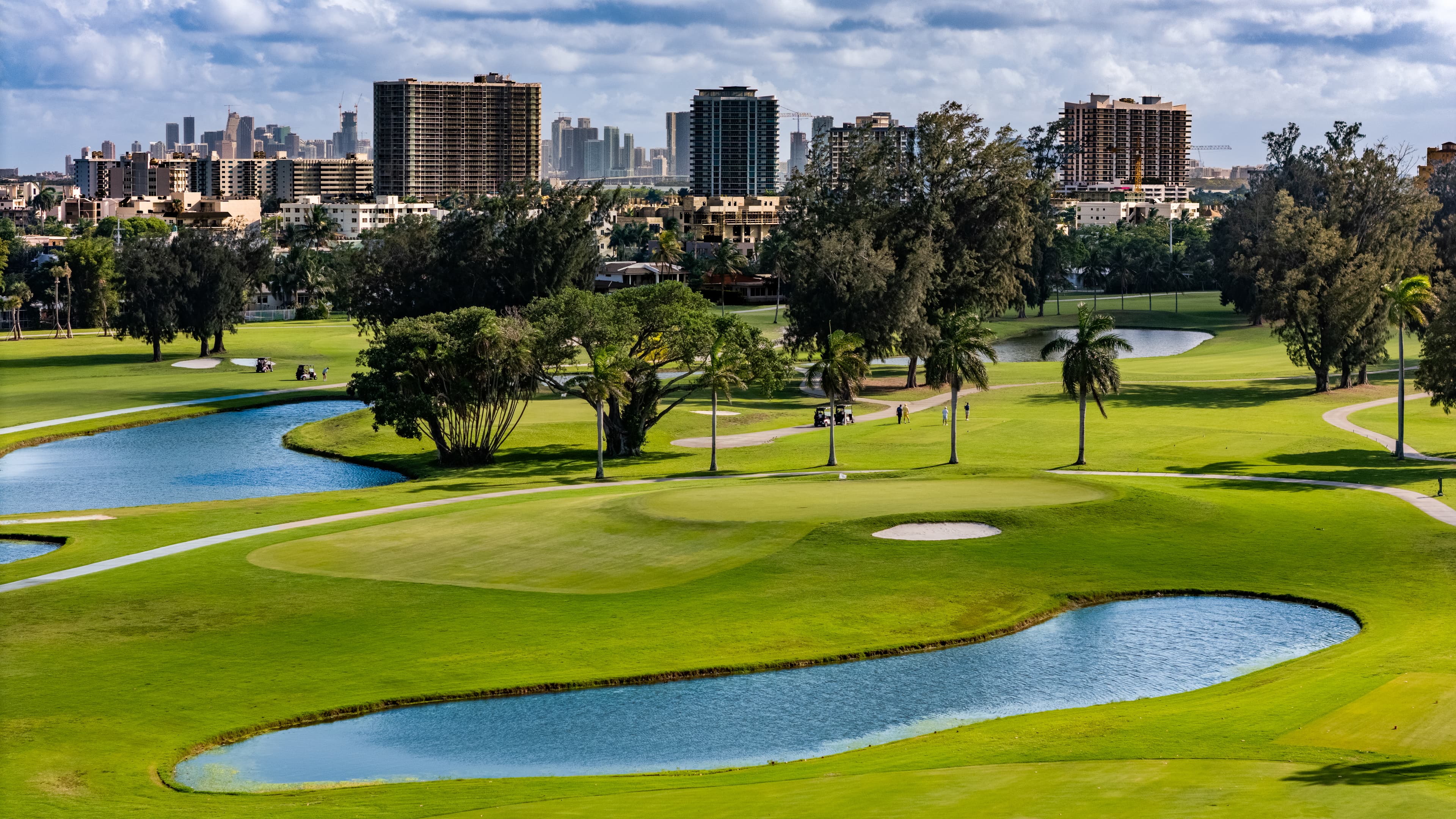 Lush green golf course with water hazards and palm trees against a city skyline.
