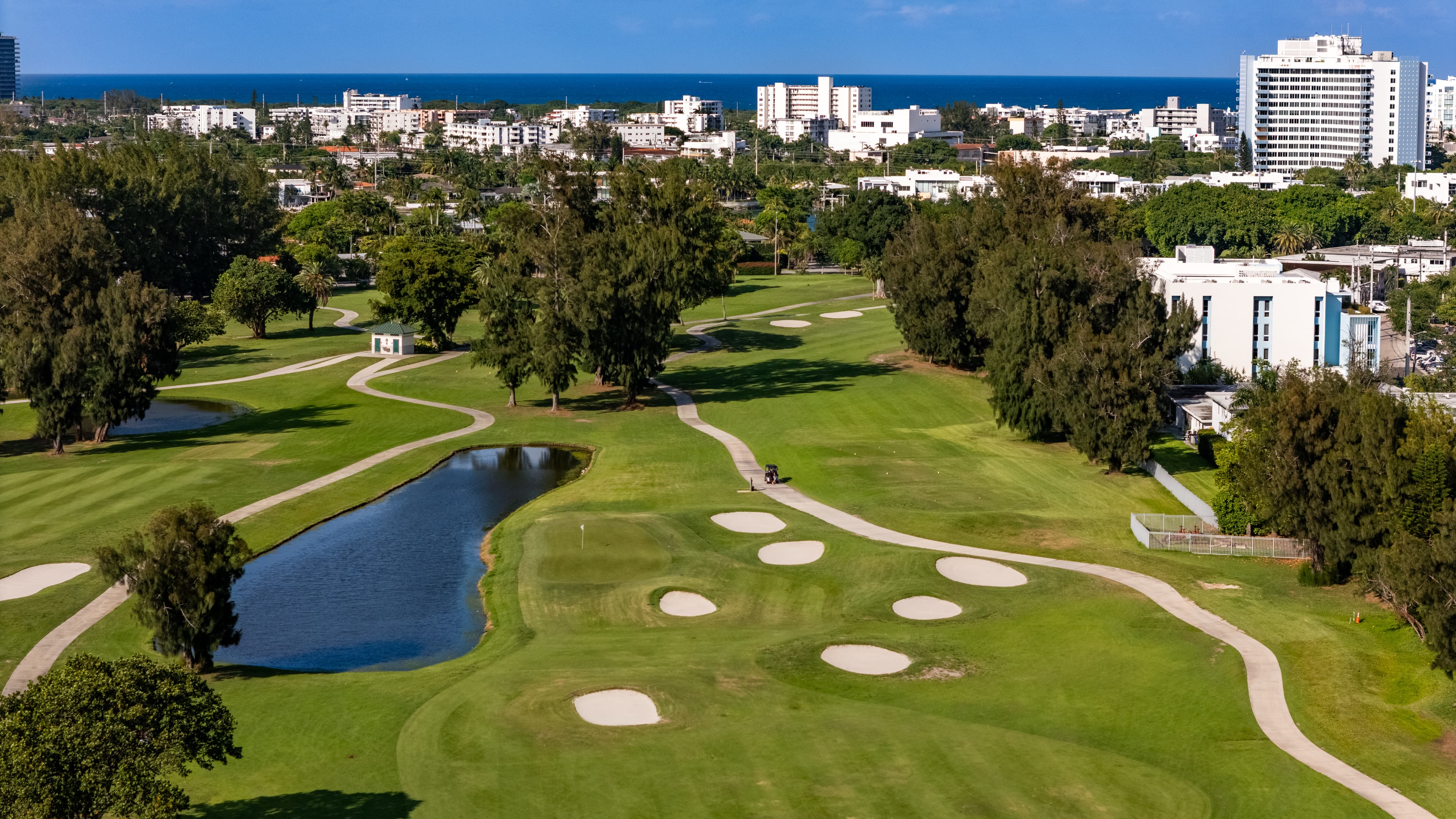 Lush green golf course with sand traps and a pond, overlooking a coastal city skyline.