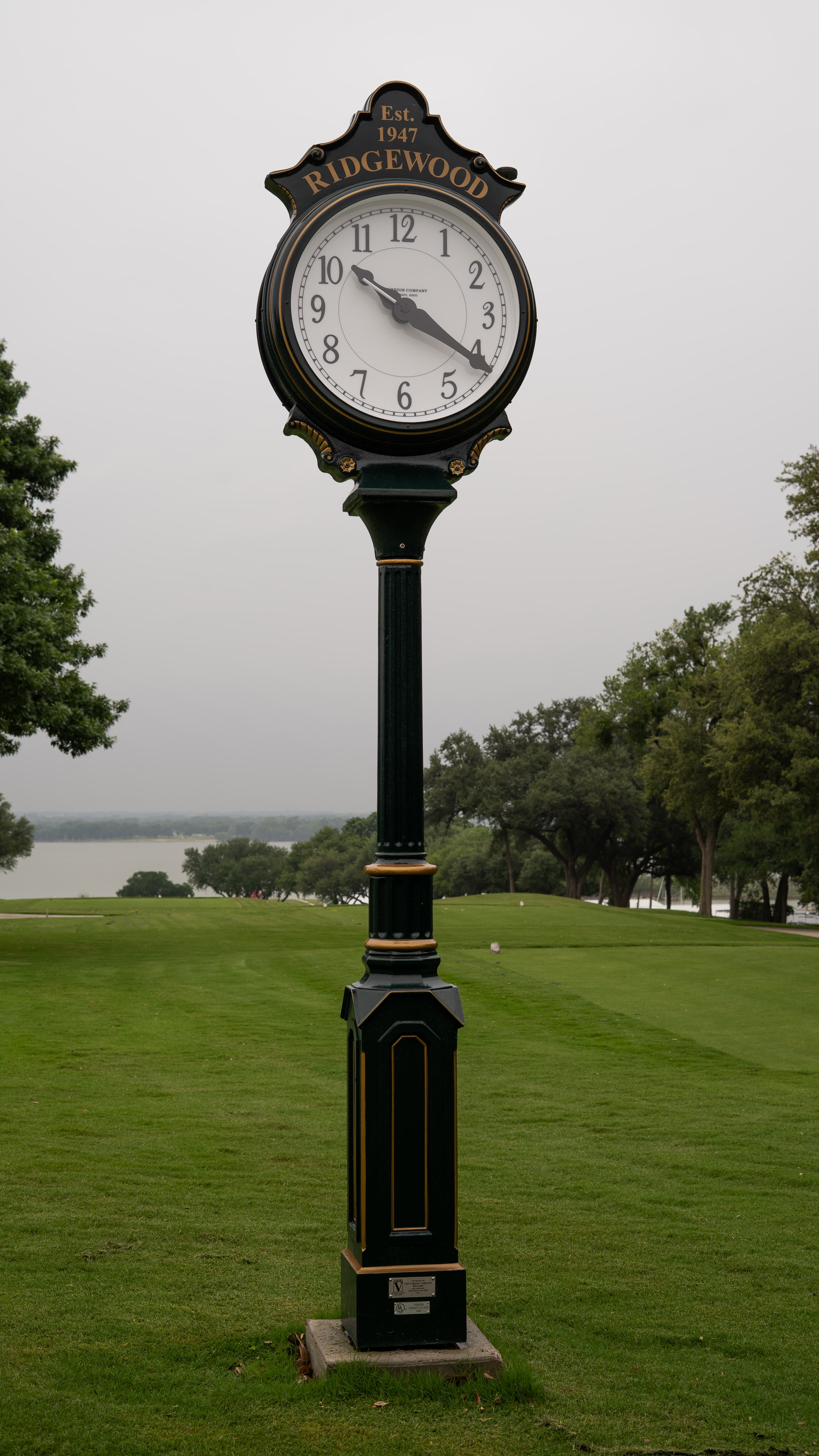 Ornate black and gold Ridgewood clock on a green lawn with trees and a lake.